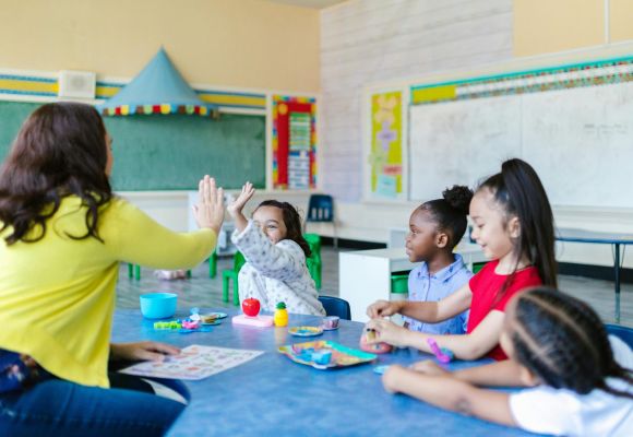 Kinder sitzen an Tisch, ein Mädchen macht High-Five mit einer Betreuerin