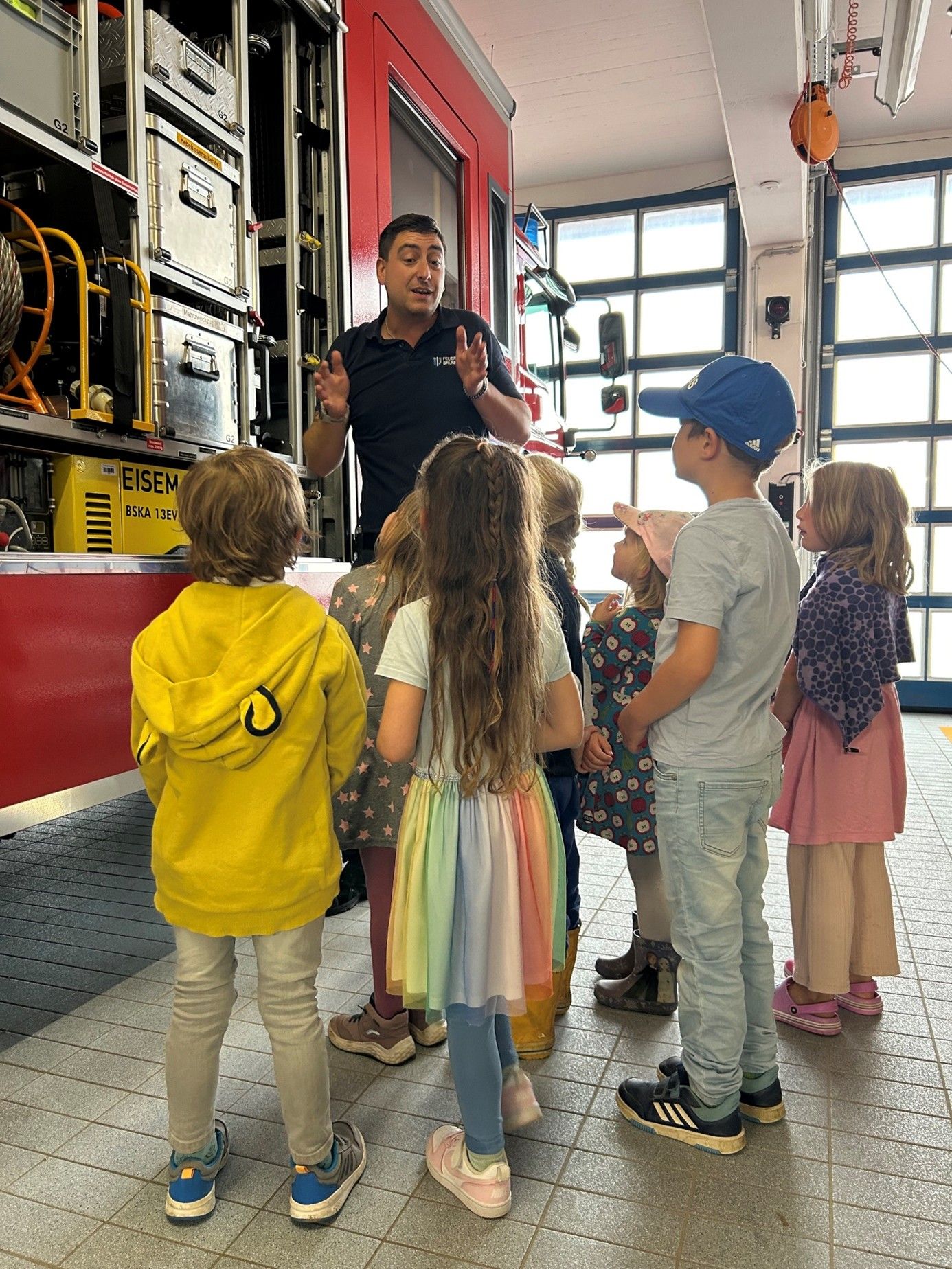 Feuerwehrmann erklärt Kindern vor Einsatzfahrzeug etwas