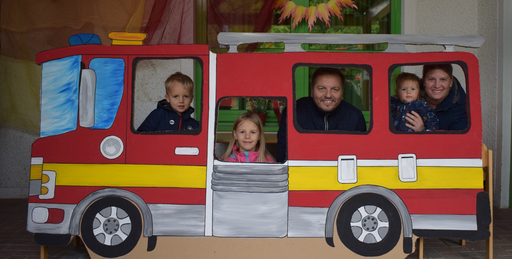 Kinder und Erwachsener hinter gemalter Feuerwehrauto-Wand