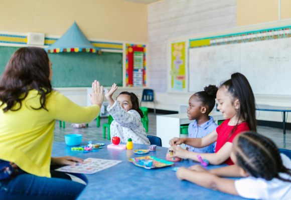 Kinder sitzen an Tisch, ein Mädchen macht High-Five mit einer Betreuerin